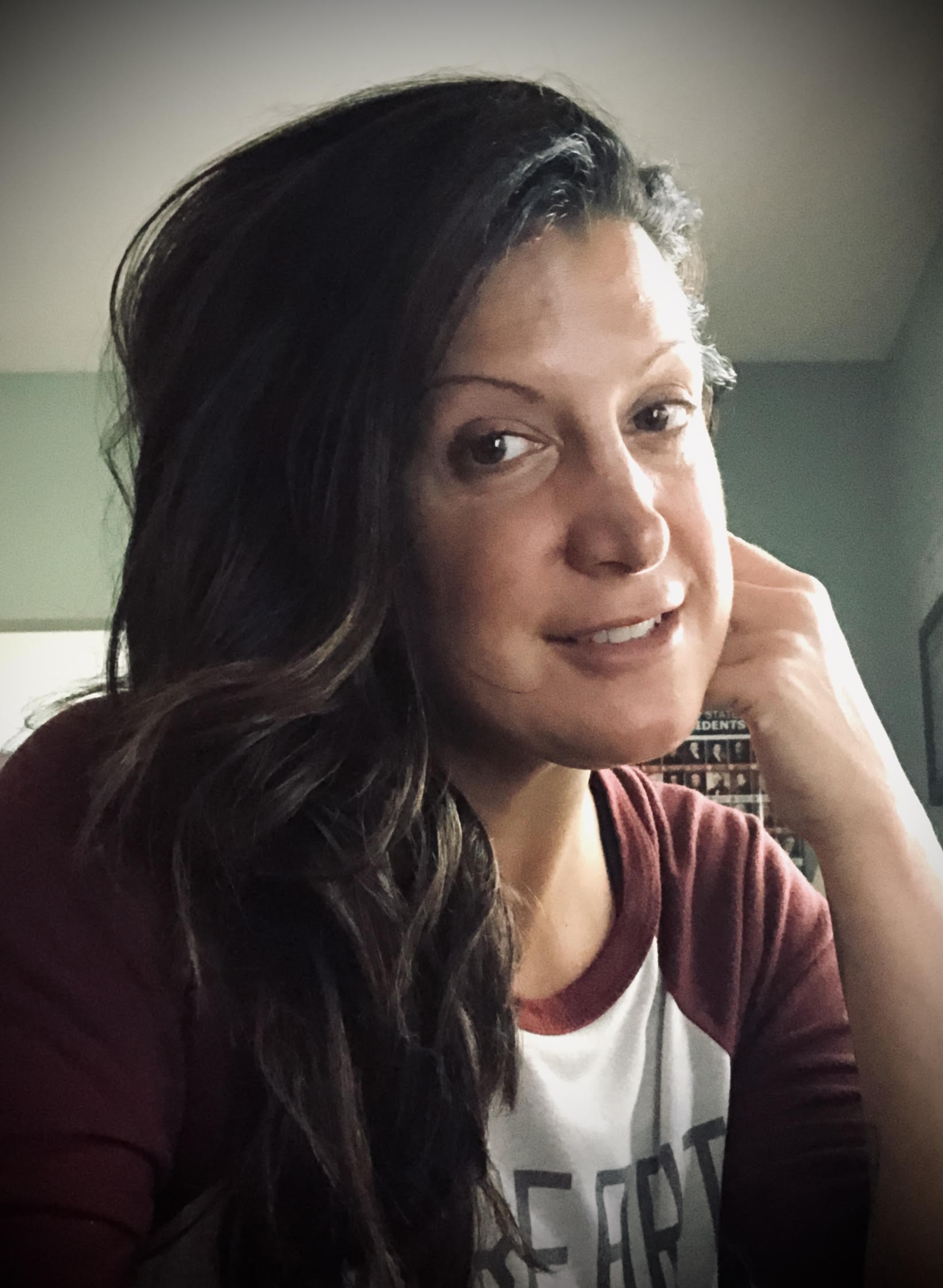 Woman with wavy hair smiling at the camera, resting her chin on her hand, wearing a striped shirt in a dimly lit room.