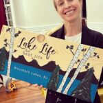 A person holding a wooden sign that reads "Lake Life, the best life" with a landscape design and "Mountain Lakes, NJ" written at the bottom.