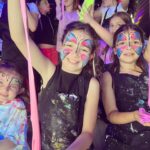 Children with colorful face paint and aprons enjoy playing with stretchy slime in a lively, playful setting.