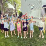 A group of children holding colorful ribbons in an outdoor setting near a house. Adults are in the background, some holding cameras. The children appear to be enjoying an activity together.