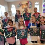 A group of nine children in aprons display their mermaid-themed art on canvas at an indoor party with pink and purple balloon decorations in the background.