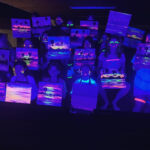 A group of people display their neon-painted canvases under black light, which makes the bright colors of their artwork and clothing glow in the dark.